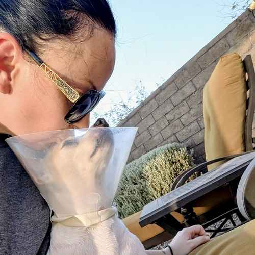 Woman cuddling a white-and-tan dog wearing a recovery cone outdoors.